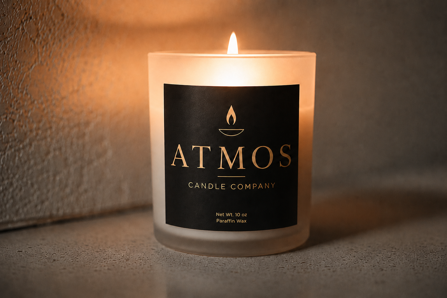 Lit ATMOS Candle Company 10 oz candle glowing on a concrete surface against a warm textured wall